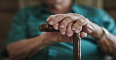 An elderly man lays both hands on his cane.