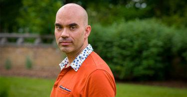 A man wearing an orange shirt peers over his left shoulder. He is standing infront of green bushes, which are blurred in the background.