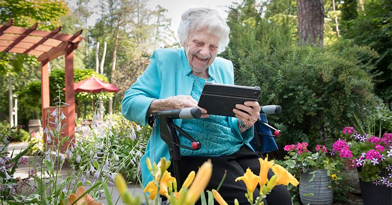 A senior woman smiles as she takes a photograph of a yellow flower.
