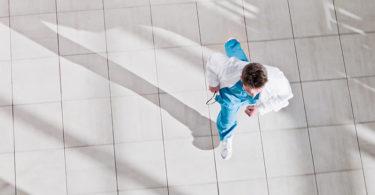 A doctor wearing light blue scrubs and a white lab coat runs down a hallway.