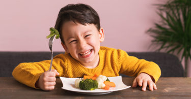 A child smiles as he lifts a piece of broccoli up with his fork.