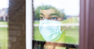 A child wearing a face mask peers out a window.