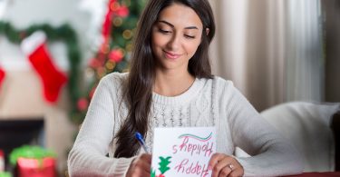 A young woman at home, surrounded by Christmas decorations, writing in a card that reads 'happy holidays'