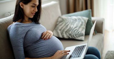 pregnant woman sits on couch looking at laptop