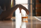 A woman's arm rests on a wooden bar counter, while her hand touches the rim of glass filled with amber-coloured liquid.