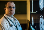 A male doctor wearing a white lab coat and stethoscope around his shoulders looks at the camera while standing next to a series of lit up brain scans