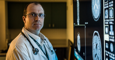 A male doctor wearing a white lab coat and stethoscope around his shoulders looks at the camera while standing next to a series of lit up brain scans