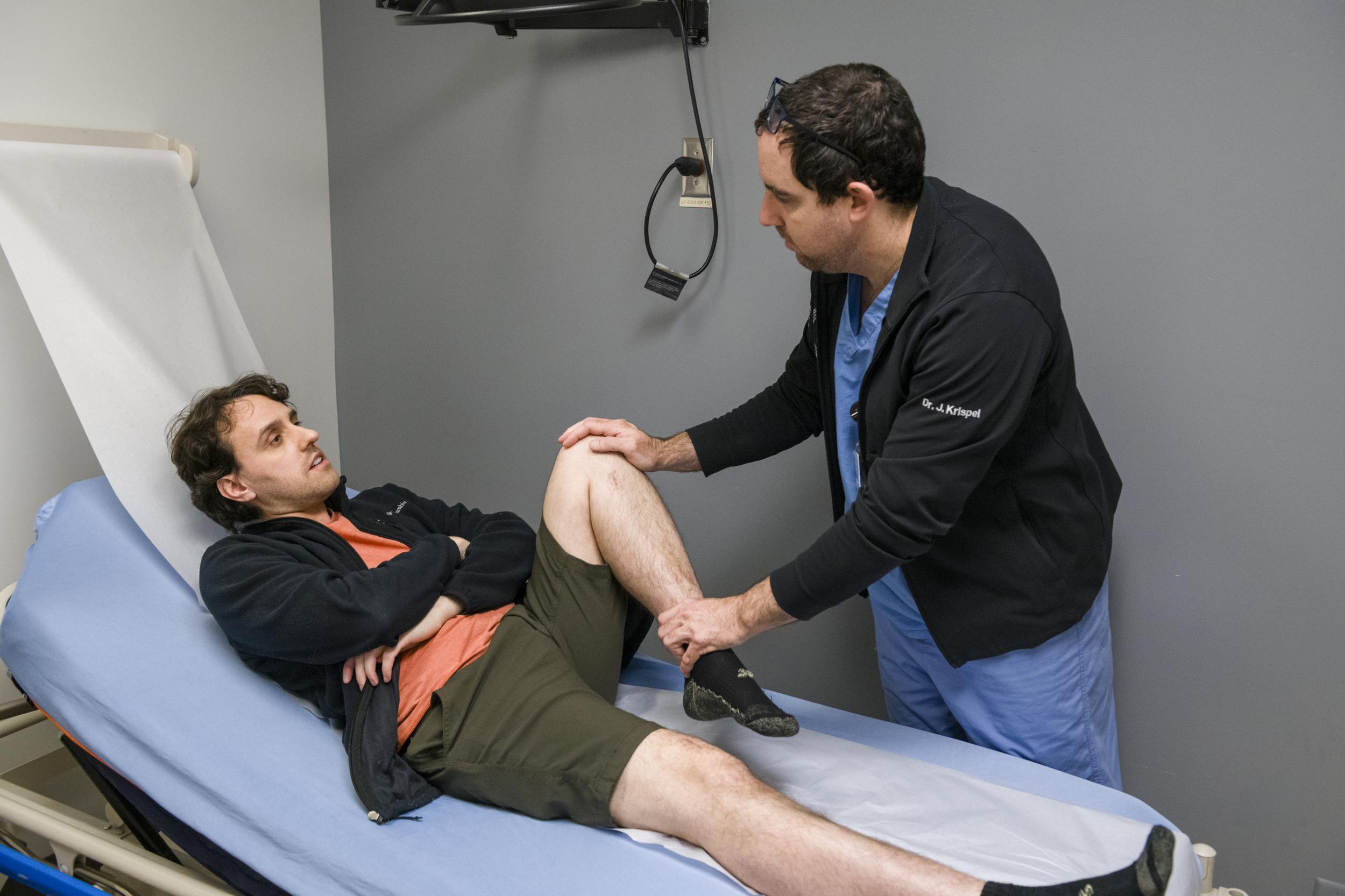 A male clinician examines a male patient's leg mobility.